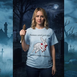 Puede incluir: Una camiseta azul claro con el texto "Bunnies aren't just cute like everybody supposes" y un gráfico de un conejo blanco con salpicaduras de sangre roja. La persona de la imagen sostiene una estaca de madera. El fondo es una escena de cementerio.
