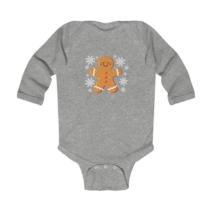 Op de afbeelding: Een grijze babybody met lange mouwen met een peperkoekenmannetje. Het peperkoekenmannetje is bruin met witte glazuurdetails en omringd door witte sneeuwvlokken. De body heeft een drukknoopsluiting aan de onderkant.