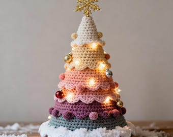 Op de afbeelding: Een handgemaakte, gehaakte kerstboom in kegelvorm, met een gradiënt van pastelkleuren. De boom is versierd met kleine ornamenten, lichtslingers en een gouden sneeuwvlok als piek. De basis is afgewerkt met wit haakwerk en staat op een houten oppervlak.
