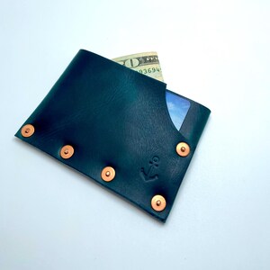 May include: Dark teal leather wallet with copper rivets and an anchor emblem. A twenty-dollar bill and a credit card are visible inside the wallet. The wallet is a minimalist design.