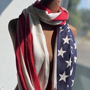 May include: An infinity scarf draped on a mannequin. The scarf features the American flag design, with red and white stripes, and a blue field with white stars. The scarf is made of a soft, lightweight fabric.