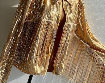 May include: A gold sequin jacket with fringe detail. The jacket has a collared neckline, a zipper closure, and a pocket on the chest. The long sleeves and body are covered in gold sequins. The jacket is displayed on a mannequin.