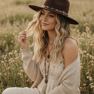 May include: A woman wearing a brown fedora hat with a decorative band, a cream-coloured embroidered dress, and a beige cardigan. She is sitting in a field of wildflowers, with long blonde hair and layered necklaces.