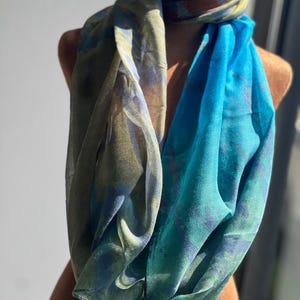 Könnte beinhalten: Ein farbenfroher Loop-Schal, drapiert auf einer Schaufensterpuppe. Der Schal zeigt eine Mischung aus Blau-, Grün- und Brauntönen. Der Stoff wirkt leicht und fließend, mit einem dezenten Muster. Ein Modeaccessoire.