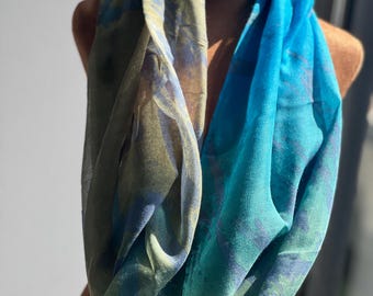 Puede incluir: Bufanda infinita multicolor, exhibida en un maniquí. La bufanda presenta una mezcla de tonos azules, verdes y marrones. La tela parece ligera y fluida, con un patrón sutil. Un accesorio de moda.