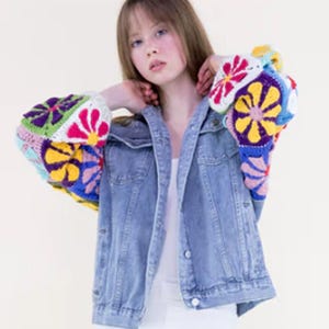 May include: A light blue denim jacket with colorful, crochet sleeves. The sleeves feature a patchwork design with flower motifs in shades of pink, yellow, green, and blue. The jacket has a classic collar and button-down front.