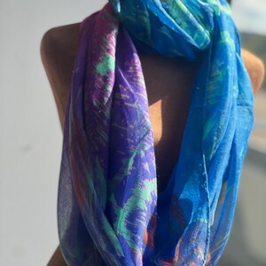 Peut inclure: Une écharpe infinie vibrante dans les tons de bleu, de violet et de vert. L'écharpe est drapée sur un mannequin, mettant en valeur son tissu fluide et léger. Le motif présente un motif semblable à une aquarelle, ajoutant une touche artistique à l'accessoire.