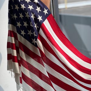 Puede incluir: Bufanda drapeada con el diseño de la bandera estadounidense. La bufanda muestra el campo azul con estrellas blancas y rayas rojas y blancas. La tela tiene una textura suave y flecos.