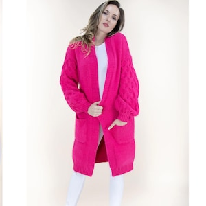 Puede incluir: Un cárdigan largo y abierto en color fucsia vibrante. El suéter presenta un diseño de punto trenzado en las mangas y dos bolsillos delanteros. La modelo lleva el cárdigan sobre una camiseta blanca y pantalones blancos.