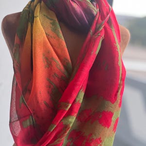 May include: A vibrant, colorful scarf with a flowing design. The scarf features a blend of red, orange, yellow, and green hues. The fabric has a semi-transparent quality, and the design appears to be a pattern of brushstrokes.