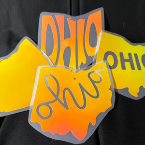 May include: Four Ohio state-shaped stickers in shades of yellow and orange, with the word "OHIO" in various fonts. The stickers have a gradient effect and a gray outline, set against a dark background.