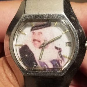 May include: A vintage silver-toned wristwatch with a rectangular face. The watch face features a faded image of a man in a dark suit and hat. The watch has a silver band and a small date window.