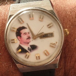 Puede incluir: Un reloj de pulsera vintage con esfera blanca con un retrato e indicadores de hora dorados. El reloj tiene una caja plateada, una correa de cuero negra y una pantalla de d&iacute;a y fecha. El reloj est&aacute; en una mu&ntilde;eca.