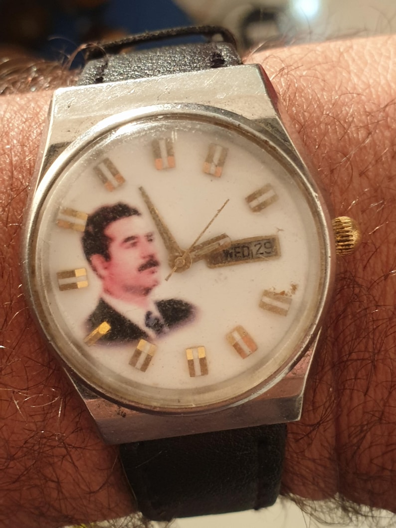 Puede incluir: Un reloj de pulsera vintage con esfera blanca con un retrato e indicadores de hora dorados. El reloj tiene una caja plateada, una correa de cuero negra y una pantalla de d&iacute;a y fecha. El reloj est&aacute; en una mu&ntilde;eca.