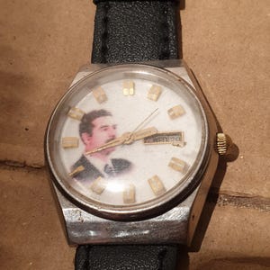 Puede incluir: Un reloj de pulsera vintage con caja plateada y correa de cuero negro. La esfera del reloj presenta un retrato e indicadores de hora dorados. El d&iacute;a y la fecha se muestran en la parte superior de la esfera.