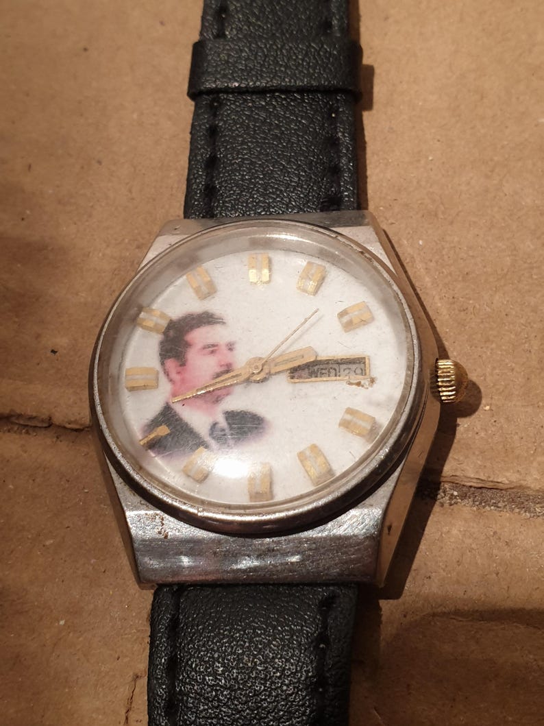 Puede incluir: Un reloj de pulsera vintage con caja plateada y correa de cuero negro. La esfera del reloj presenta un retrato e indicadores de hora dorados. El d&iacute;a y la fecha se muestran en la parte superior de la esfera.