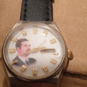 Puede incluir: Un reloj de pulsera vintage con caja plateada y correa de cuero negro. La esfera del reloj presenta un retrato e indicadores de hora dorados. El d&iacute;a y la fecha se muestran a las 3 en punto.