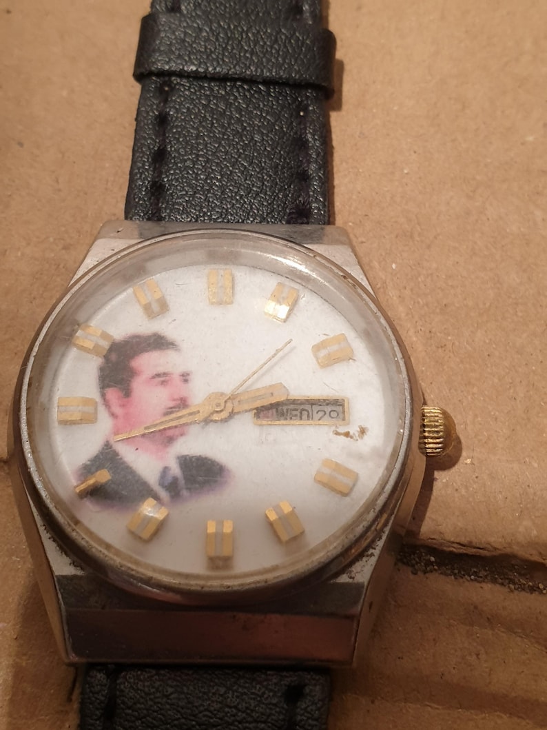 Puede incluir: Un reloj de pulsera vintage con caja plateada y correa de cuero negro. La esfera del reloj presenta un retrato e indicadores de hora dorados. El d&iacute;a y la fecha se muestran a las 3 en punto.