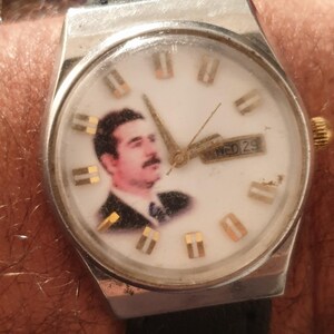 Puede incluir: Un reloj de pulsera vintage con caja plateada y correa de cuero negro. La esfera blanca del reloj presenta un retrato e indicadores de hora dorados. El d&iacute;a y la fecha se muestran a las 3 en punto.