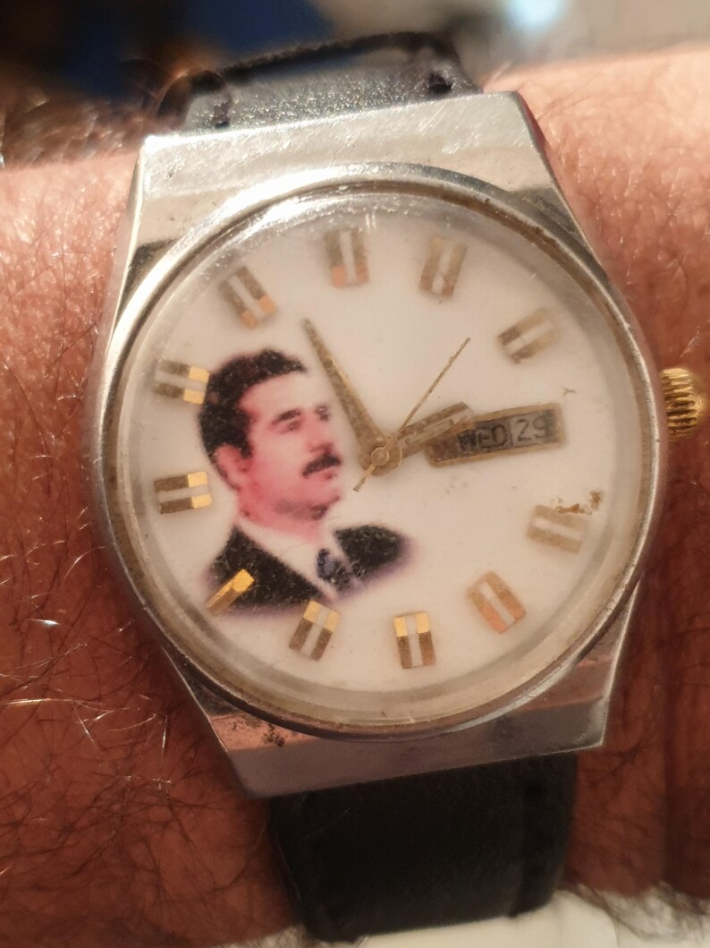 Puede incluir: Un reloj de pulsera vintage con caja plateada y correa de cuero negro. La esfera blanca del reloj presenta un retrato e indicadores de hora dorados. El d&iacute;a y la fecha se muestran a las 3 en punto.