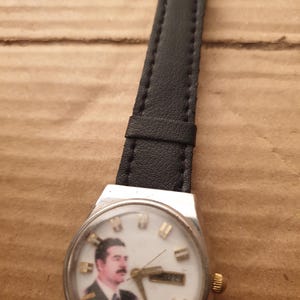 Puede incluir: Un reloj de pulsera vintage con caja plateada y correa de cuero negro. La esfera del reloj presenta un retrato de un hombre con bigote. Las manecillas indican la hora y la fecha es visible.