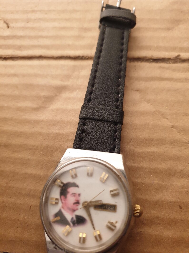 Puede incluir: Un reloj de pulsera vintage con caja plateada y correa de cuero negro. La esfera del reloj presenta un retrato de un hombre con bigote. Las manecillas indican la hora y la fecha es visible.