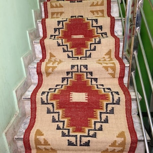 Op de afbeelding: Een traploper met een geometrisch ontwerp in rood, zwart en goud op een beige achtergrond. Het tapijt is gemaakt van een geweven materiaal en heeft een franje aan de onderkant. Het tapijt ligt op marmeren trappen.