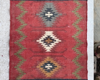 May include: A rectangular woven rug featuring a red background with green zigzag side borders and three central diamond-shaped geometric patterns in grey, white, and brown. The rug includes grey end panels with colourful motifs and fringe.