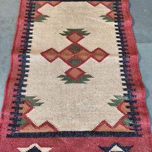 Op de afbeelding: Een rechthoekig jute vloerkleed met een beige middenveld en een rode rand. De rand heeft geometrische patronen in marine, bruin en groen. Het ontwerp van het vloerkleed bevat ruit- en zigzagmotieven.