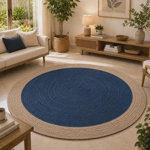 Puede incluir: Una alfombra trenzada circular grande que presenta un centro azul marino sólido rodeado por un borde ancho de color beige natural. La alfombra está construida con fibras texturizadas estrechamente enrolladas, creando un patrón concéntrico uniforme.