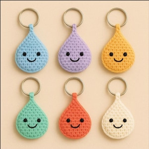 May include: Six handmade crochet teardrop keychains in various colors: light blue, purple, yellow, green, orange, and cream. Each keychain features a smiling face and a gold-colored metal ring.