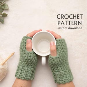 Op de afbeelding: Een paar saliegroene gehaakte vingerloze handschoenen die een witte mok vasthouden. De afbeelding bevat een bol garen, een haaknaald en de tekst "CROCHET PATTERN instant download".