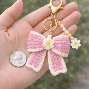 May include: A handmade pink crochet bow keychain with a white daisy center and trim. The keychain has a gold-colored clasp and a beaded accent with a small white daisy charm. A quarter is next to the bow.