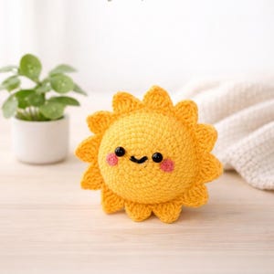 Puede incluir: Un sol amarillo tejido a ganchillo con una cara sonriente y mejillas rosadas. El sol tiene un centro redondo y rayos puntiagudos. El texto "CozyLoopStudio" está en el fondo. El sol está sobre una superficie de madera clara.