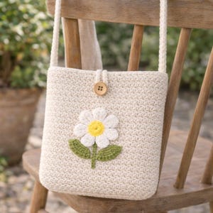 May include: A cream-coloured, crocheted shoulder bag with a large daisy appliqué. The daisy has white petals, a yellow centre, and green leaves. The bag has a button closure and is hanging on a wooden chair.