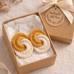 May include: A pair of handmade crochet earrings in a gift box. The earrings have a spiral design in mustard yellow and cream. The brown gift box has a cream ribbon and a tag that reads "handmade with Love".