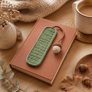 May include: A rectangular green crochet bookmark with a textured stitch pattern. Attached to the top is a thin brown cord ending in a small, three-dimensional crocheted acorn charm featuring a brown cap and a light beige, rounded base.