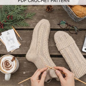 Puede incluir: Primer plano de calcetines de punto beige con diseño de trenzas. La imagen incluye agujas de tejer, lana y una taza de café, sugiriendo un proyecto artesanal. El texto dice "INSTANT DOWNLOAD PDF CROCHET PATTERN".