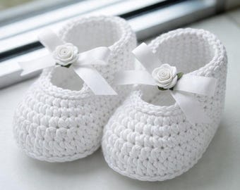 May include: A pair of white crochet baby shoes. Each shoe features a white ribbon and a small white rose accent. The shoes are designed with a Mary Jane style, perfect for a newborn.