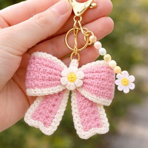 May include: A pink crochet bow keyring with a white trim and a small daisy in the centre. The keyring has a gold-coloured clasp and a beaded accent with pastel-coloured beads.