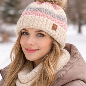 Könnte beinhalten: Eine cremefarbene Strickmütze mit rosa und grauen Streifen und einem Kunstfell-Bommel. Die Mütze hat einen kleinen braunen Aufnäher mit einem Herzsymbol. Ein passender cremefarbener Schal ist ebenfalls zu sehen. Das Wintermützen- und Schal-Set.