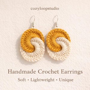May include: A pair of handmade crochet earrings. Each earring features a spiral design with a mustard yellow top section and a cream-colored bottom section. The earrings are suspended from silver-colored hooks. The text "cozyloopstudio" is at the top.