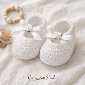 May include: A pair of white crochet baby shoes with a Mary Jane style. Each shoe features a white ribbon and a small white rose. A wooden bead bracelet and a pacifier are also in the image. The text "CozyLoop Studio" is at the bottom.