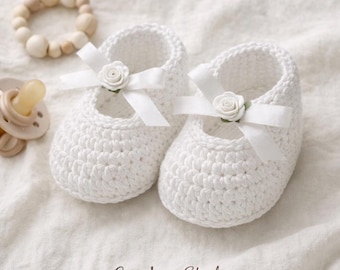 May include: A pair of white crochet baby shoes with a Mary Jane style. Each shoe features a white ribbon and a small white rose. A wooden bead bracelet and a pacifier are also in the image. The text "CozyLoop Studio" is at the bottom.