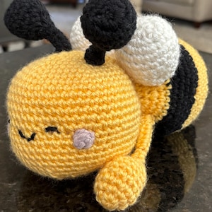 May include: A handmade crochet bee plushie. The bee features a yellow body with black stripes, black antennae, and white and black eyes. The bee has a small purple cheek and a smiling face.