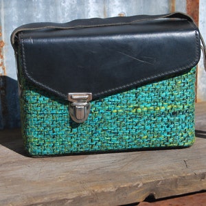 May include: A vintage camera bag with a black leather flap and a green and blue woven fabric body. The bag has a silver metal clasp.