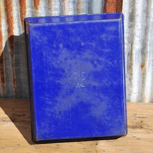 May include: A rectangular, cobalt blue tray with a slightly textured surface. The tray is set against a weathered, corrugated metal background and a wooden surface. The tray is empty.