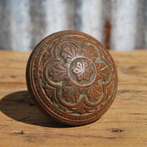 May include: A round, antique bronze door knob with an ornate floral design. The knob is aged with a patina, showing shades of brown and green. It sits on a weathered wooden surface, suggesting a vintage aesthetic.