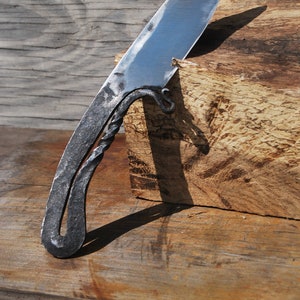K&ouml;nnte beinhalten: Ein handgeschmiedetes Messer mit einem gedrehten Griff und einer scharfen Klinge. Das Messer liegt auf einem St&uuml;ck Holz.