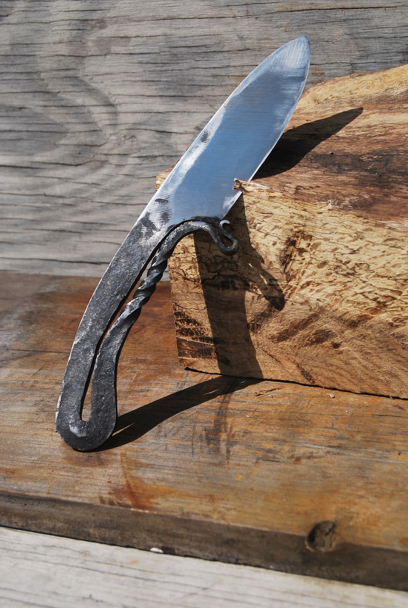 K&ouml;nnte beinhalten: Ein handgeschmiedetes Messer mit einem gedrehten Griff und einer scharfen Klinge. Das Messer liegt auf einem St&uuml;ck Holz.